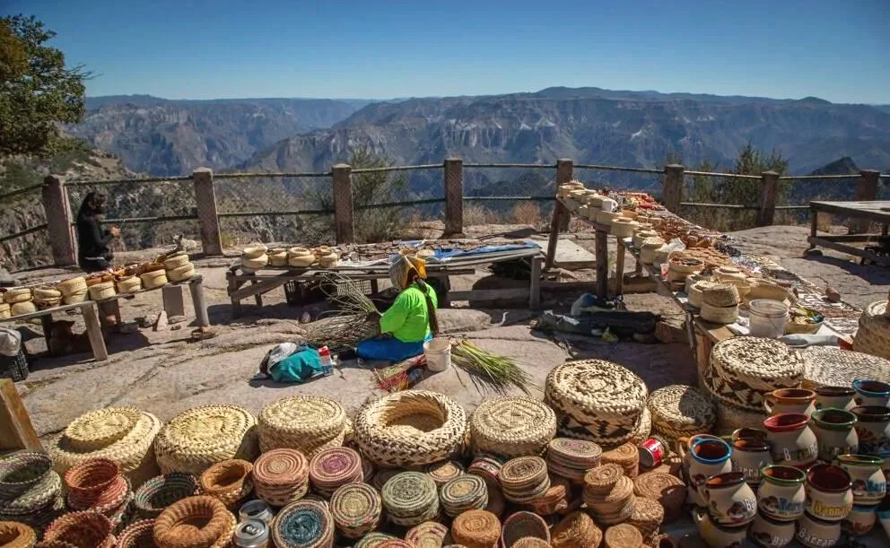 Outdoor marketplace set on a cliff overlooking a canyon with pottery and woven baskets for sale, two vendors and a person taking photos.