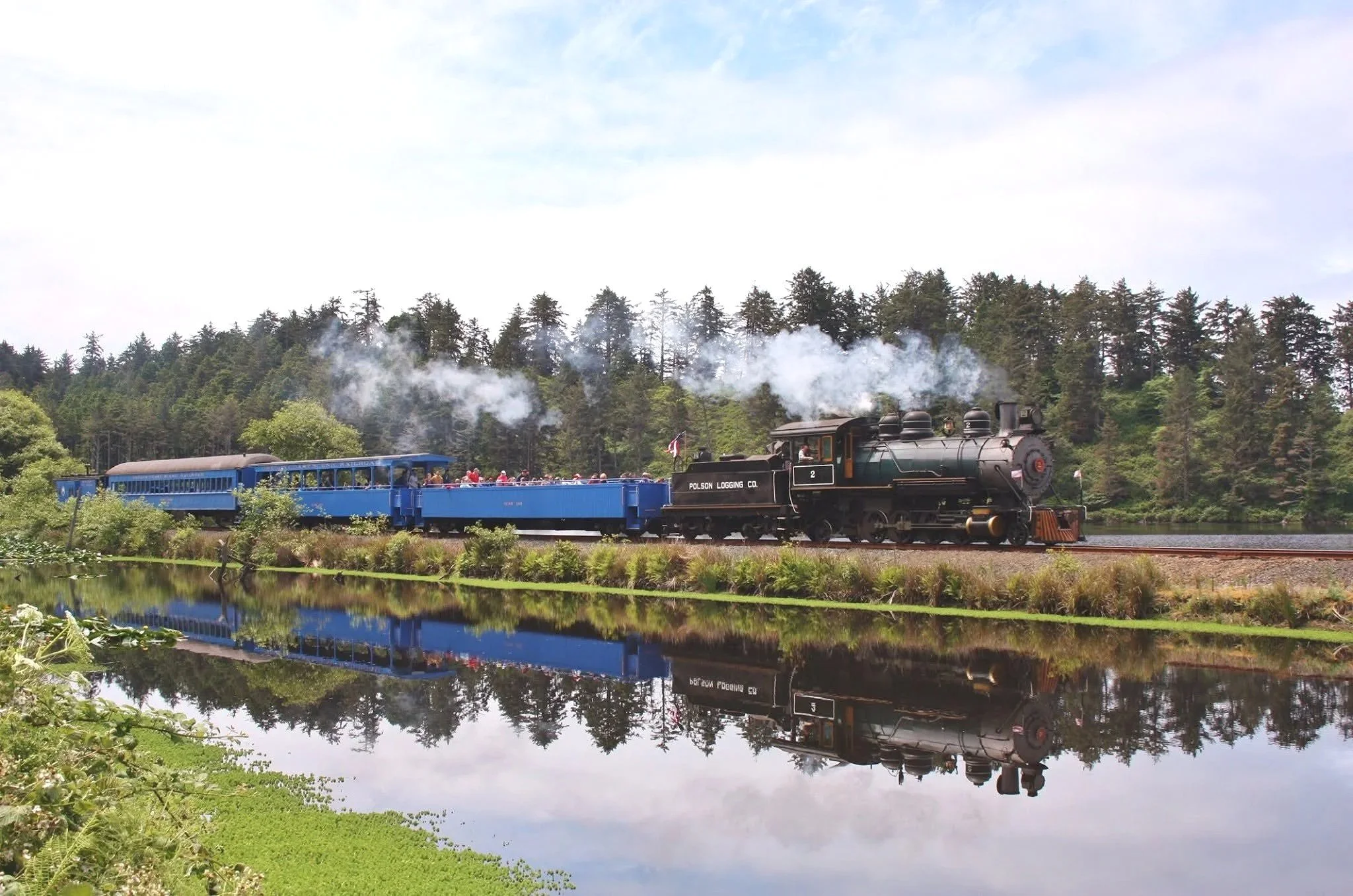 A vintage steam locomotive pulling a blue passenger train along a scenic track through a lush green landscape with a pond in the foreground, and a forested hill in the background, under a partly cloudy sky.