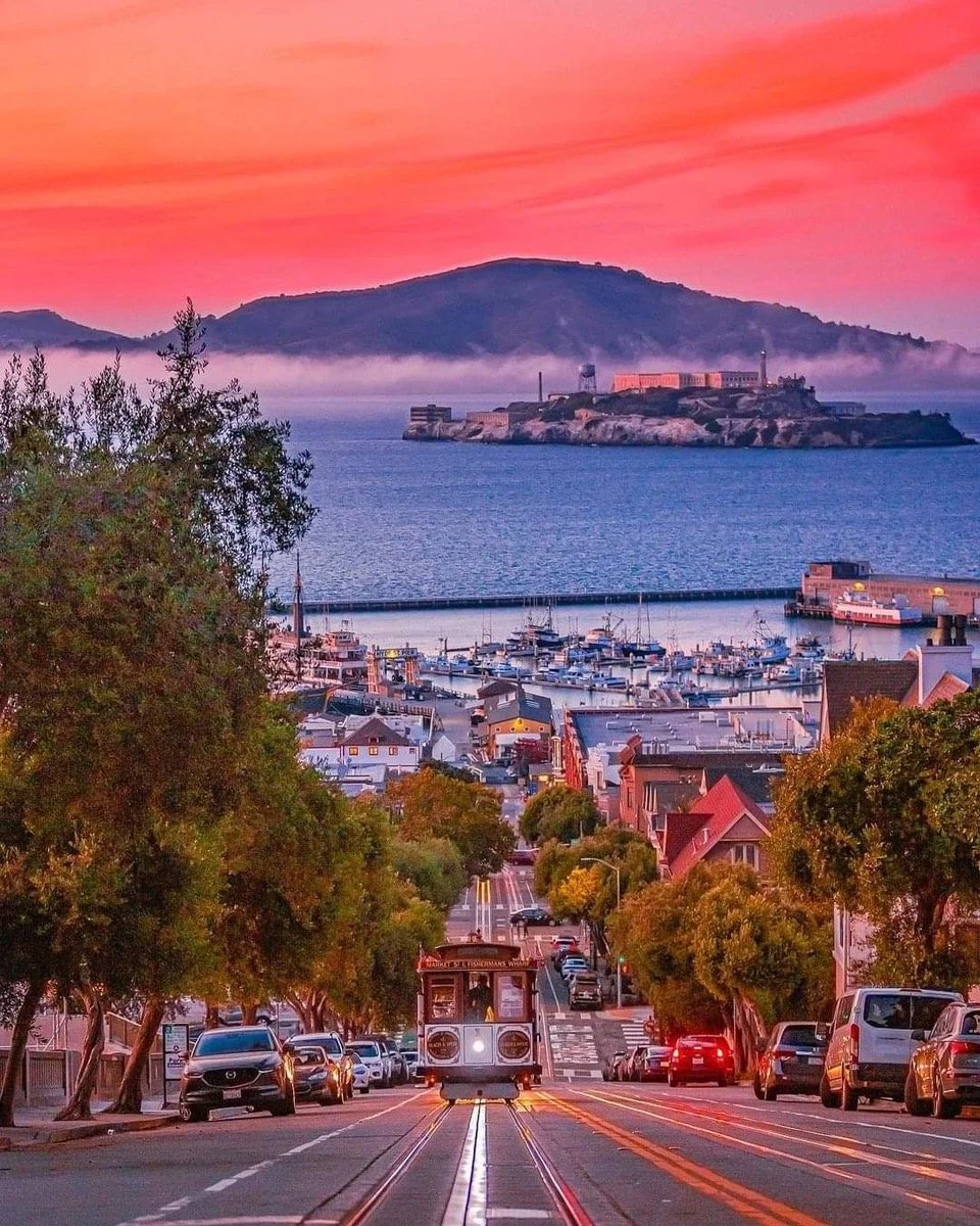 A sunset over a city with a trolley on tracks leading downhill, overlooking a harbor with boats, a bay, and Alcatraz Island in the distance, with hills and colorful clouds in the sky.