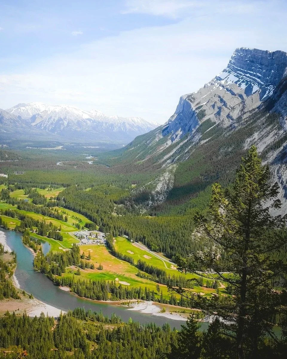 A scenic view of a mountain valley with a river winding through a lush green landscape, surrounded by snow-capped mountains and dense forests under a partly cloudy sky.