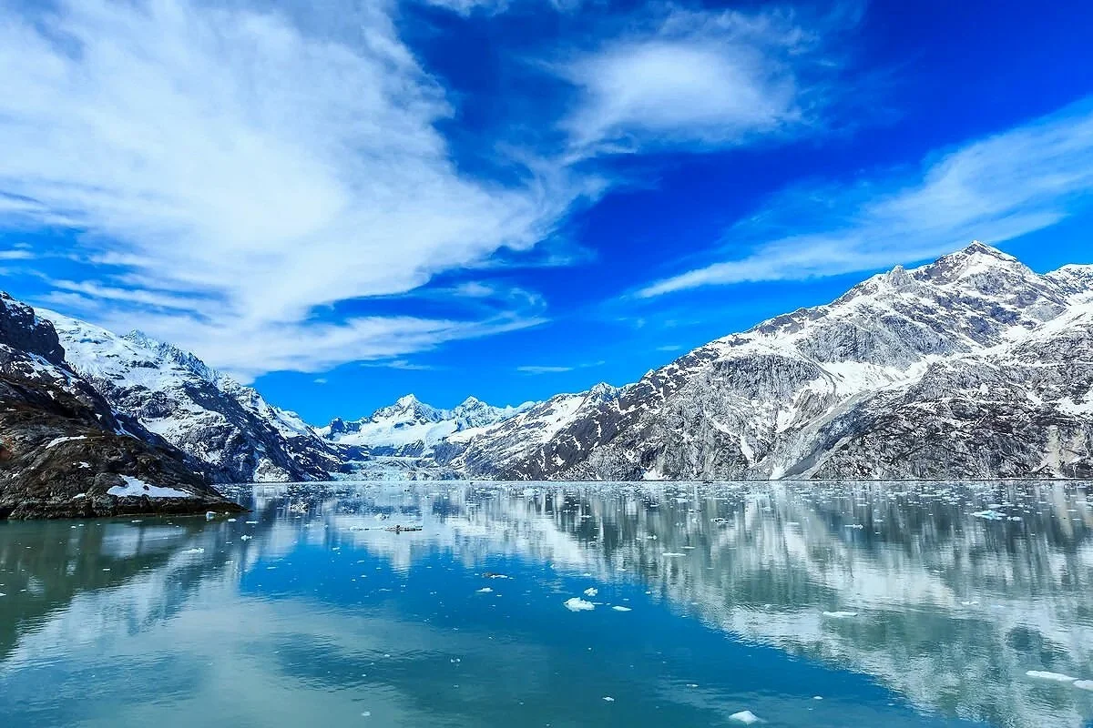 Snow-capped mountains beside a calm, icy lake under a partly cloudy blue sky.