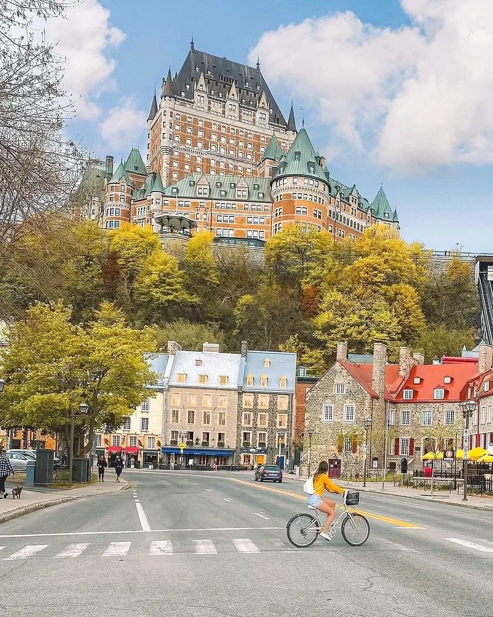 A woman riding a bicycle across a city street with historic buildings. A large castle-like hotel is visible on a hilltop in the background, with trees in the foreground.