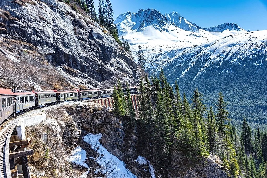 A scenic mountain landscape with a train traveling along a track on the side of a mountain, snow-capped peaks in the distance, and a forest of pine trees in the foreground.
