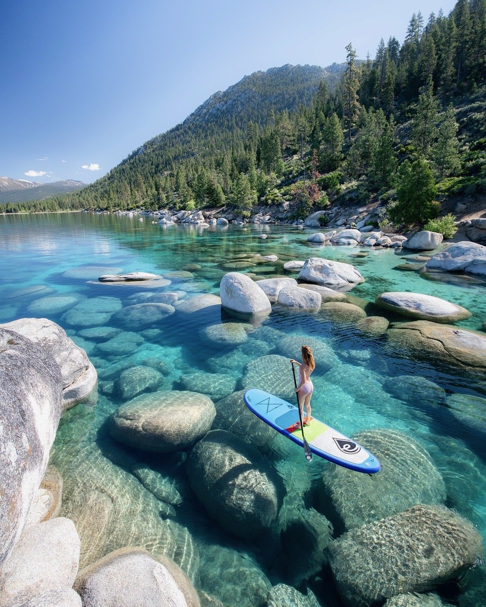 A woman paddleboarding on a clear, rocky lake with a forested mountain in the background.