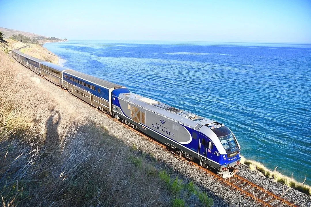 A Pacific Surfliner train traveling along a coastal railway with ocean views and cliffs in the background.