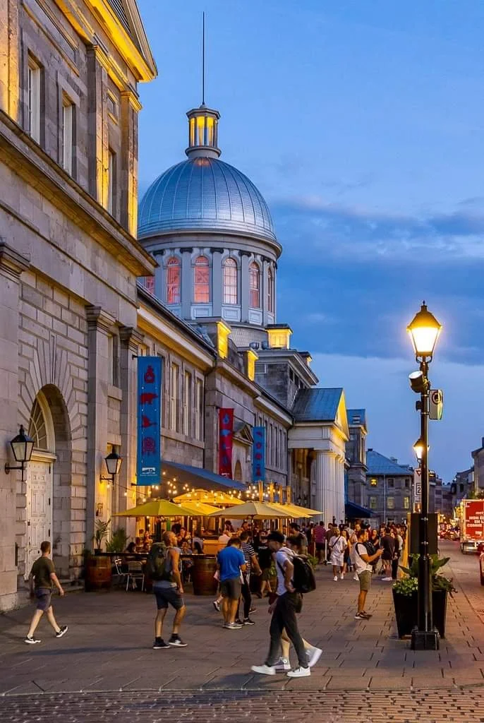 Street scene outside a historic building with a large dome, during early evening, with people walking and sitting at outdoor tables under yellow umbrellas, illuminated street lamps, and a cloudy sky.