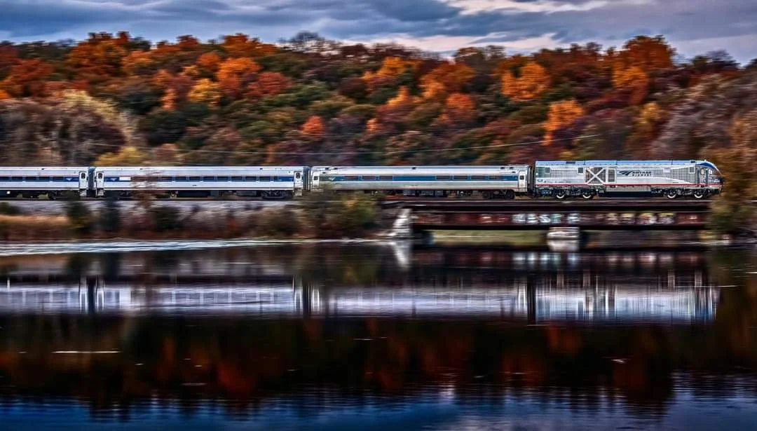 A sleek modern train traveling across a bridge over a calm body of water, with autumn-colored trees and hills in the background.