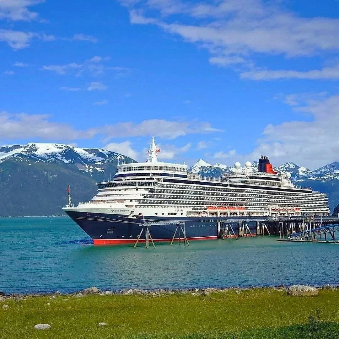 Passenger cruise ship docked at a port with snow-capped mountains in the background.