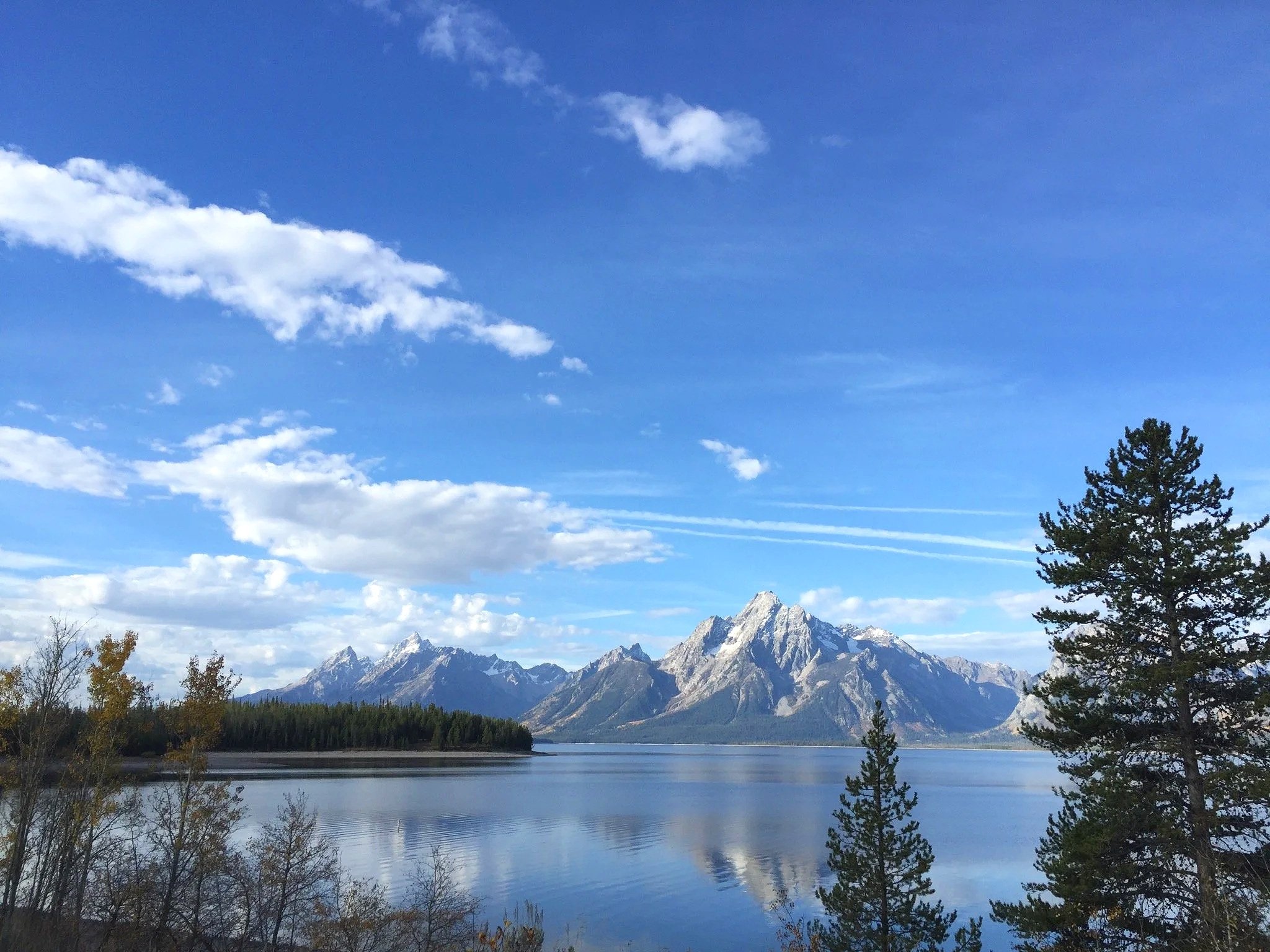 Scenic view of a lake with calm water, surrounded by trees, mountains in the background, and a partly cloudy blue sky.