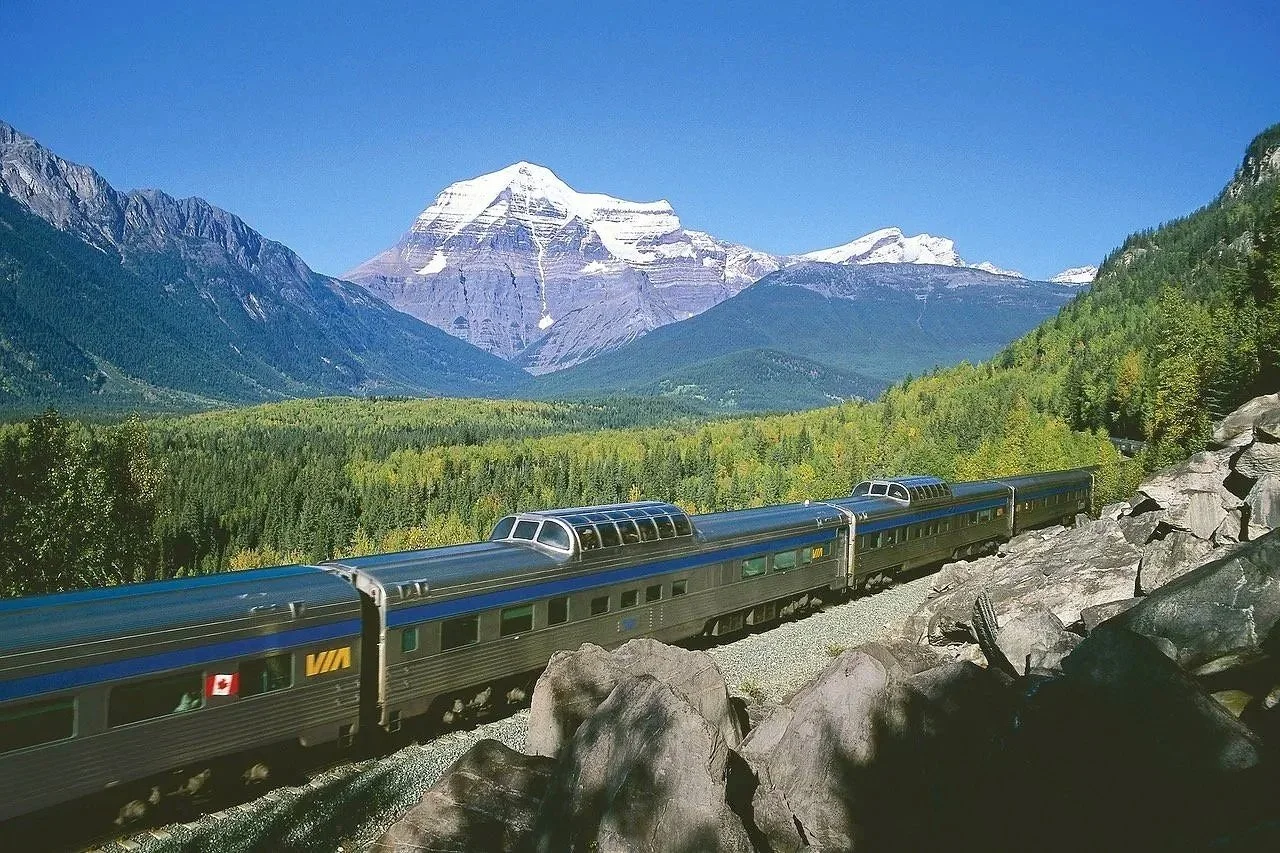 A luxury train traveling through a scenic mountain landscape with snow-capped peaks, dense green forests, and a clear blue sky.