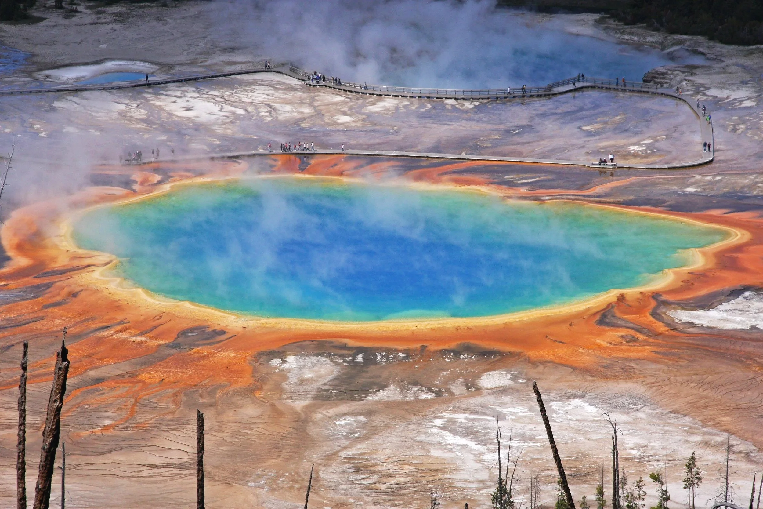 Hot spring with colorful mineral deposits and steam rising, surrounded by a boardwalk with visitors walking and observing the area.