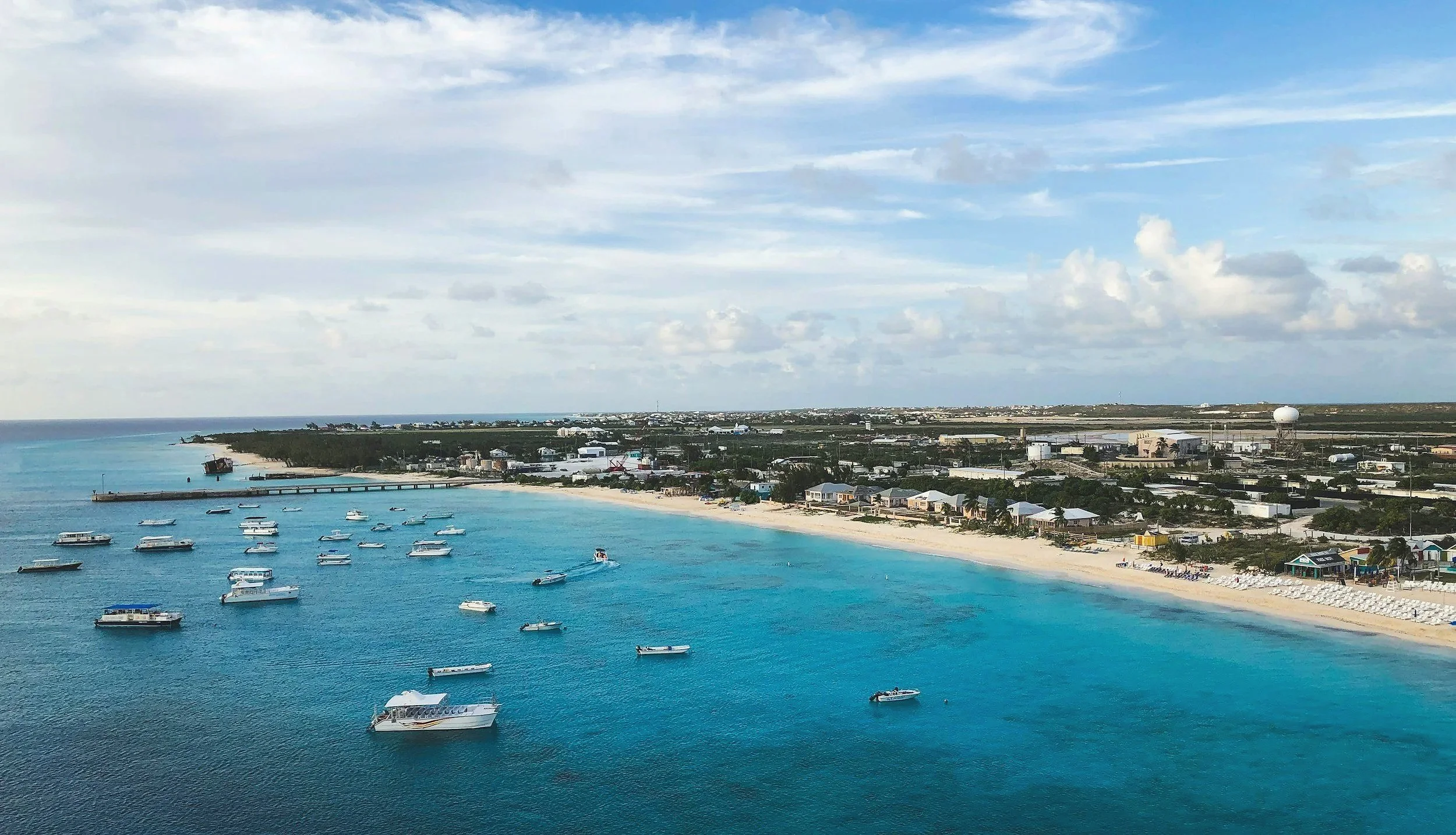 A coastal scene with clear blue waters, a sandy beach, boats anchored in the water, and a small town with buildings and infrastructure along the shoreline under a partly cloudy sky.