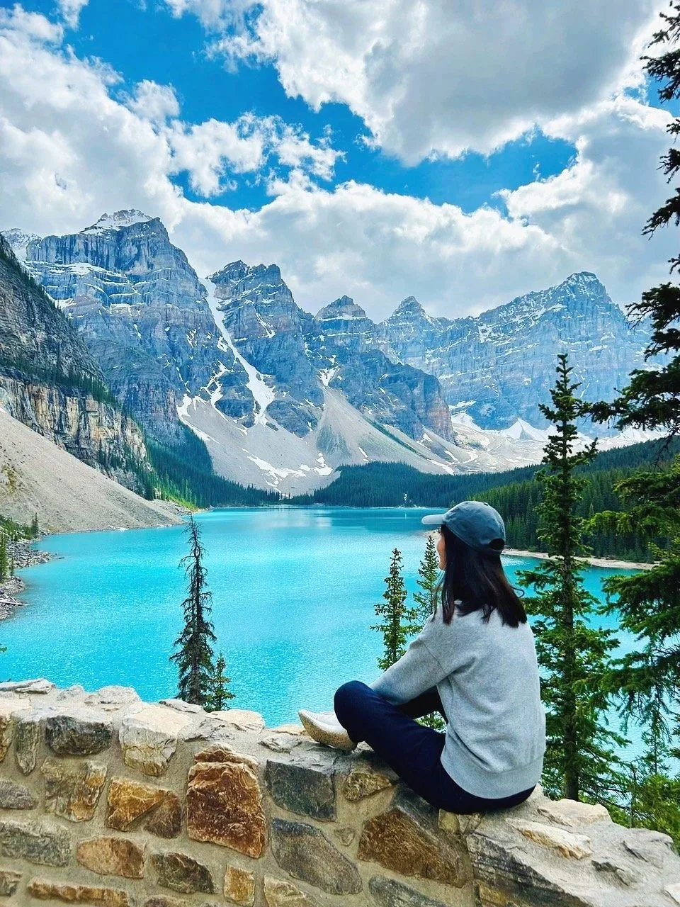 A woman with a gray cap and sweatshirt sitting on a stone ledge overlooking a bright blue lake, surrounded by evergreen trees and snow-capped mountains under a partly cloudy sky.