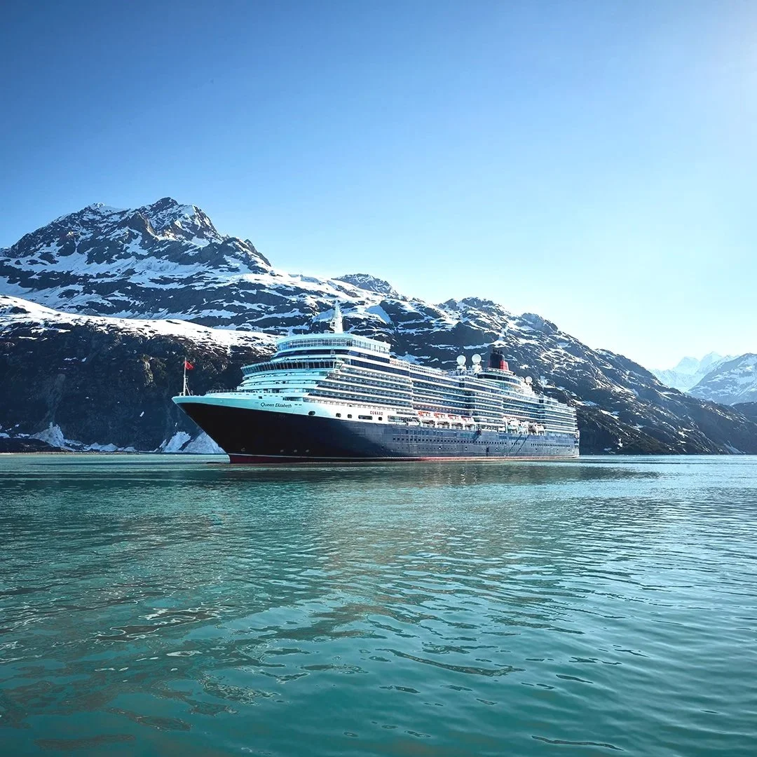 A large cruise ship sailing in calm water near snow-covered mountains under a clear blue sky.