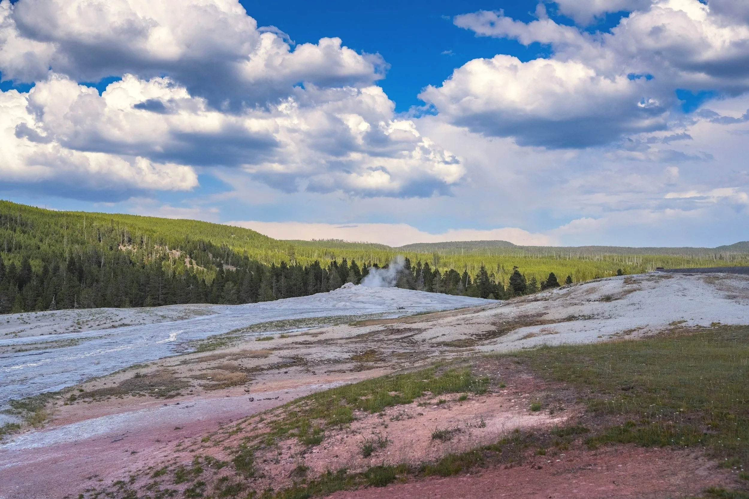 A landscape of green forested hills under a blue sky with scattered white clouds, and a geothermal area with steaming ground in the foreground.