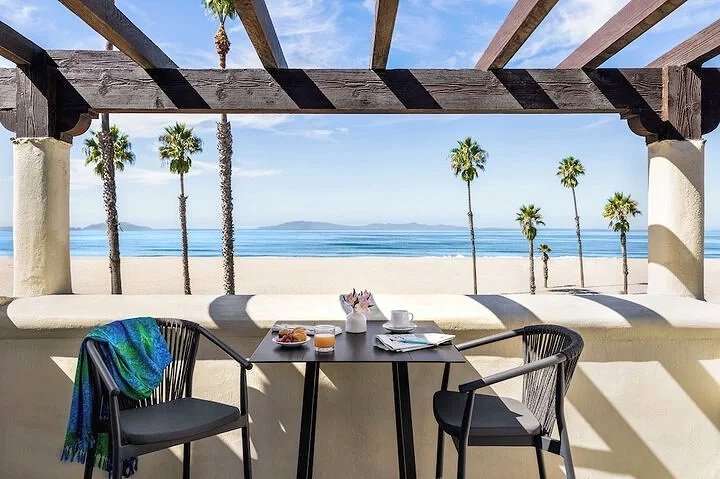 Breakfast table setup on a balcony with a view of the beach, palm trees, and ocean, under a wooden pergola.