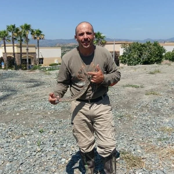 A man standing on rocky terrain outdoors, holding a small alligator, with palm trees and buildings in the background.