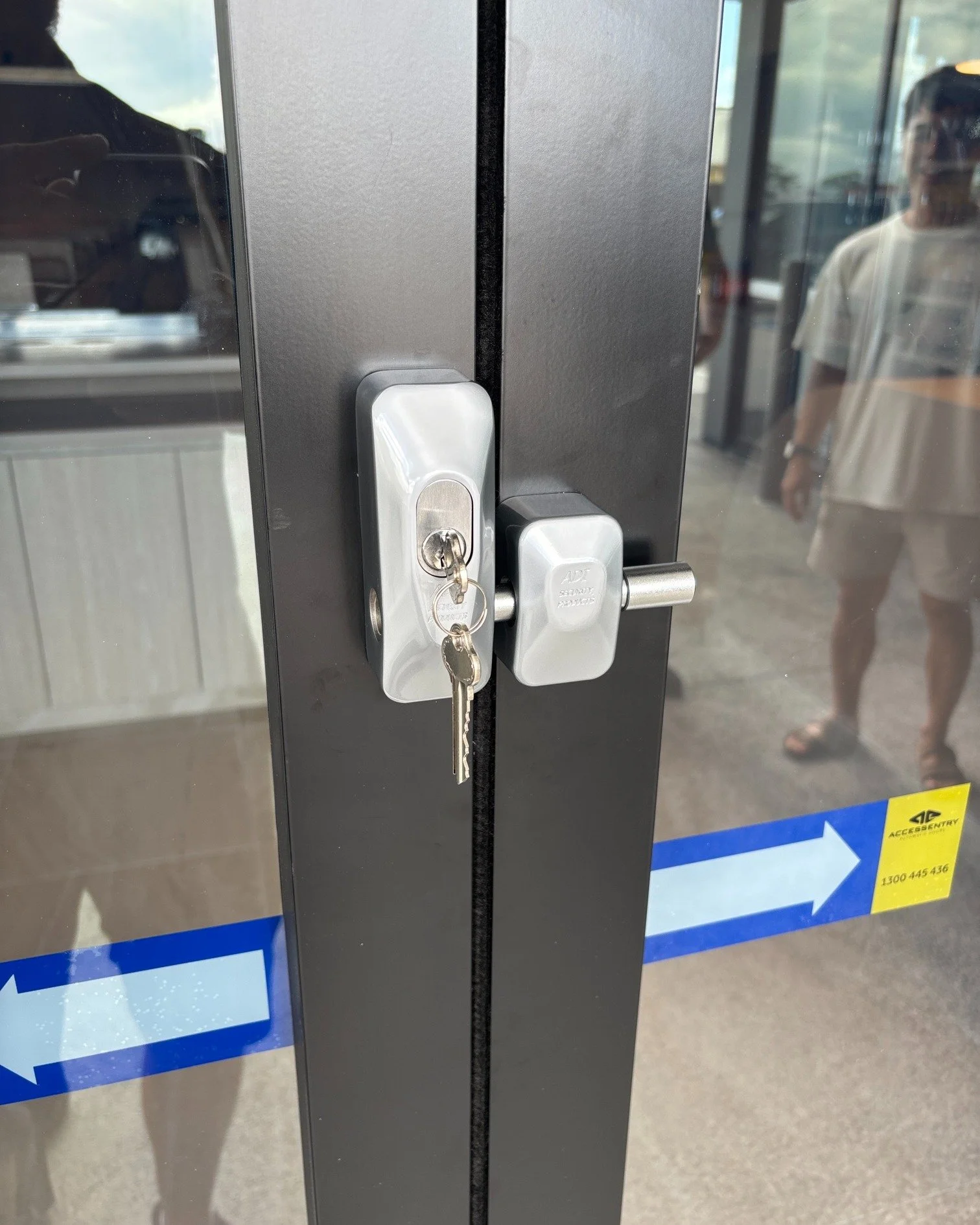 Close-up of a glass door with a silver lock and handle, with a key inserted into the lock. Reflective surfaces show people and a blue and yellow sign in the background.