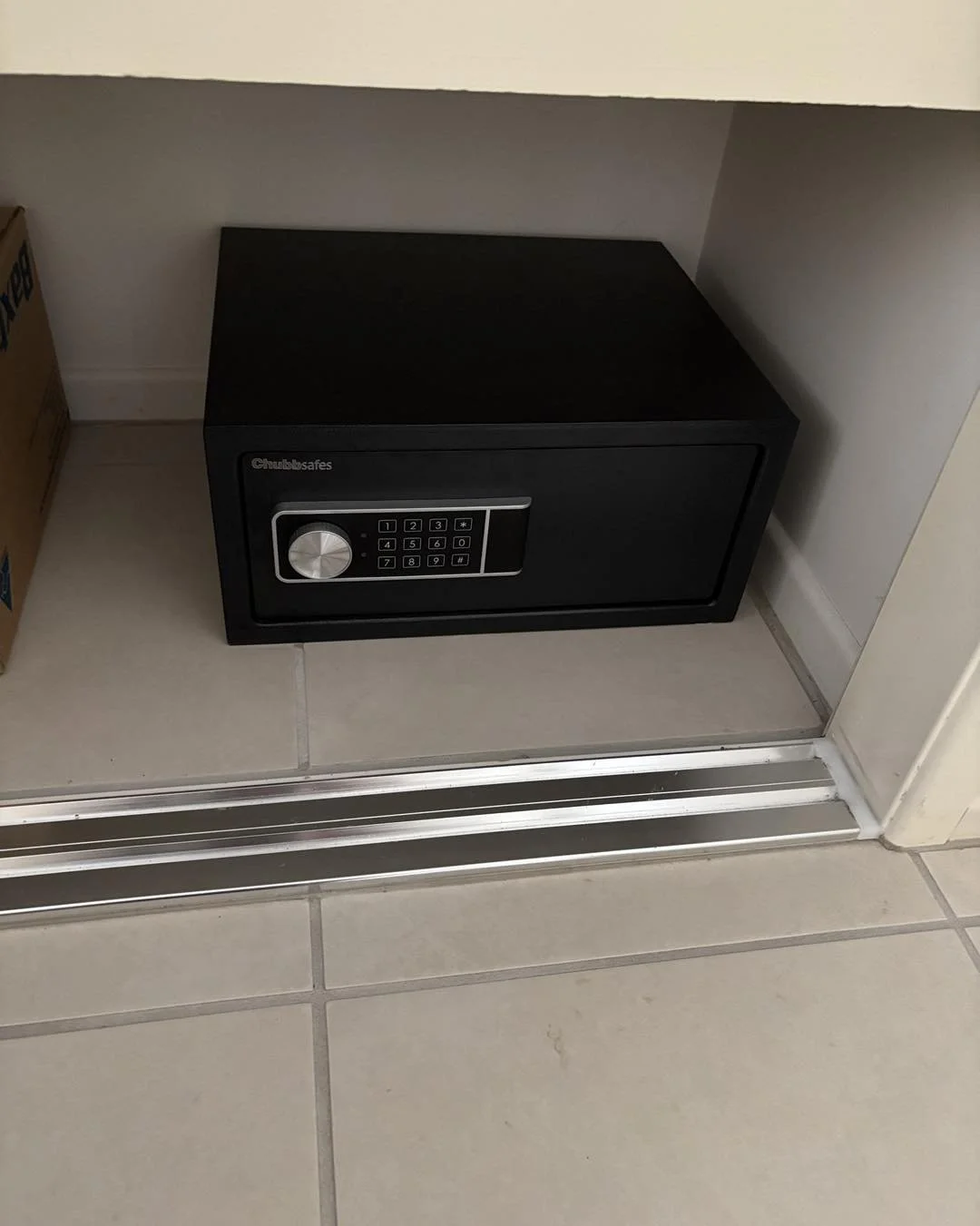 A black safe with digital keypad and lock, placed on a beige tiled floor next to a cardboard box and a white wall.