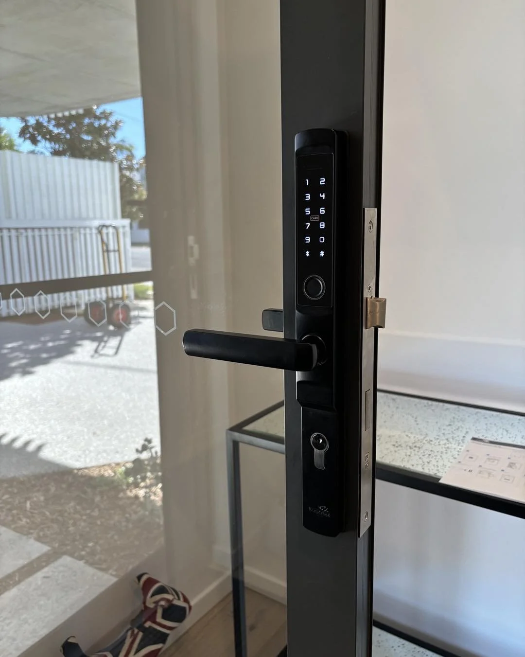 Digital keypad door lock on a glass door with a handle, leading outside. Outdoor area visible through the glass door.