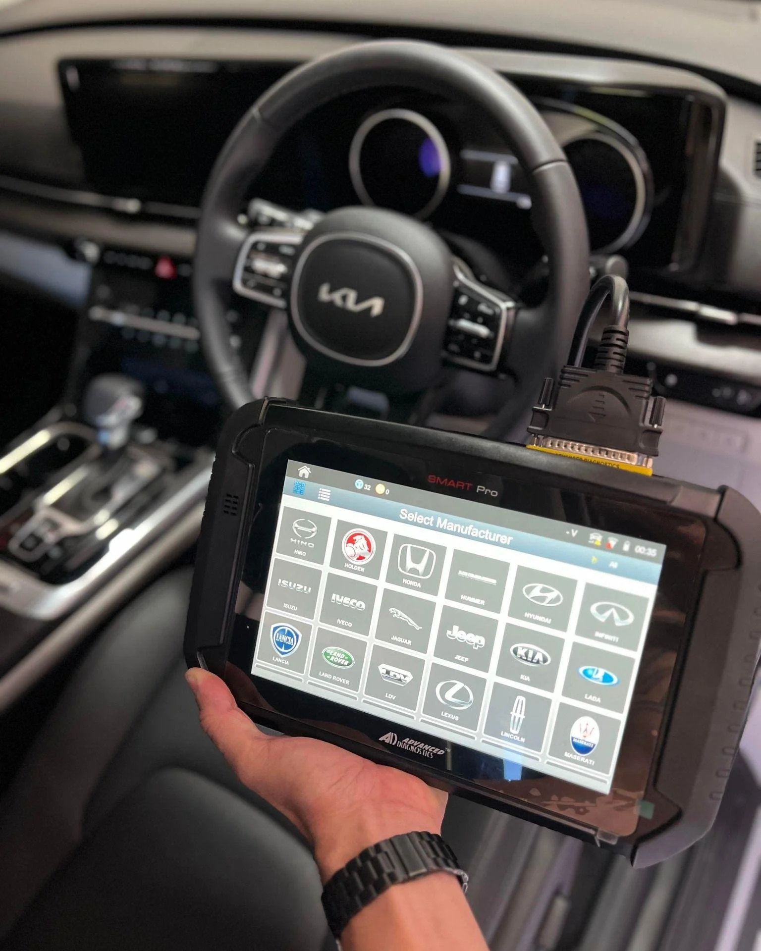 A diagnostic scan tool displaying car manufacturer logos, held inside a vehicle with a Kia steering wheel in the background.