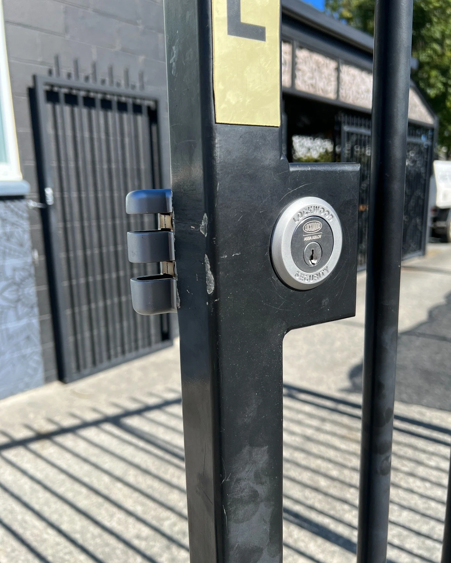 Close-up of a black lockable gate latch with a silver lock that has the brand name 'Lockwood' on it. The gate is outdoors, with a sidewalk and a building in the background.