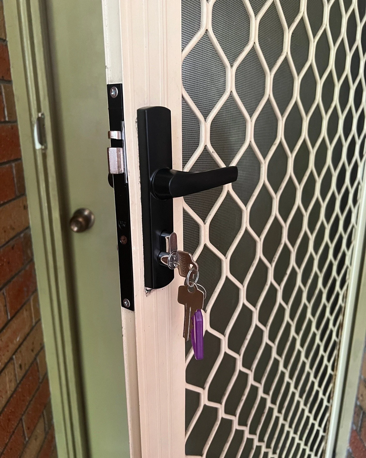 Close-up of a door lock with keys hanging on the handle, set in a door with a wire mesh screen.