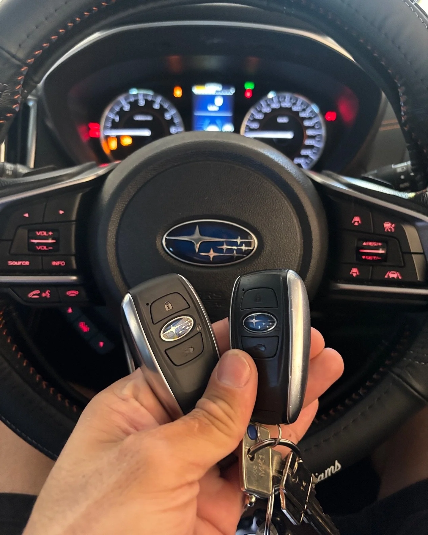A person's hand holding two Subaru car key fobs in front of a Subaru vehicle's steering wheel and dashboard.