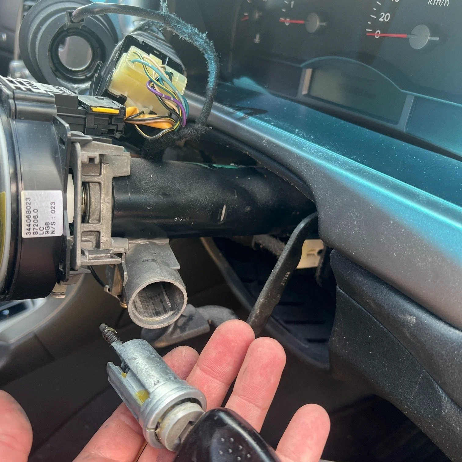 A person holding a car ignition switch removed from the steering column of a vehicle, with exposed wiring and dashboard visible.
