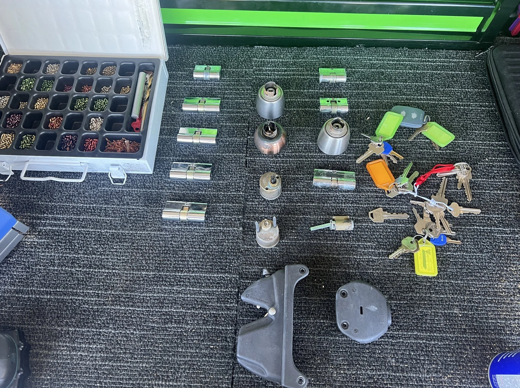 A collection of keys, key fobs, locks, and lock parts laid out on a black textured surface, with a box of small colored fasteners in a cubby.