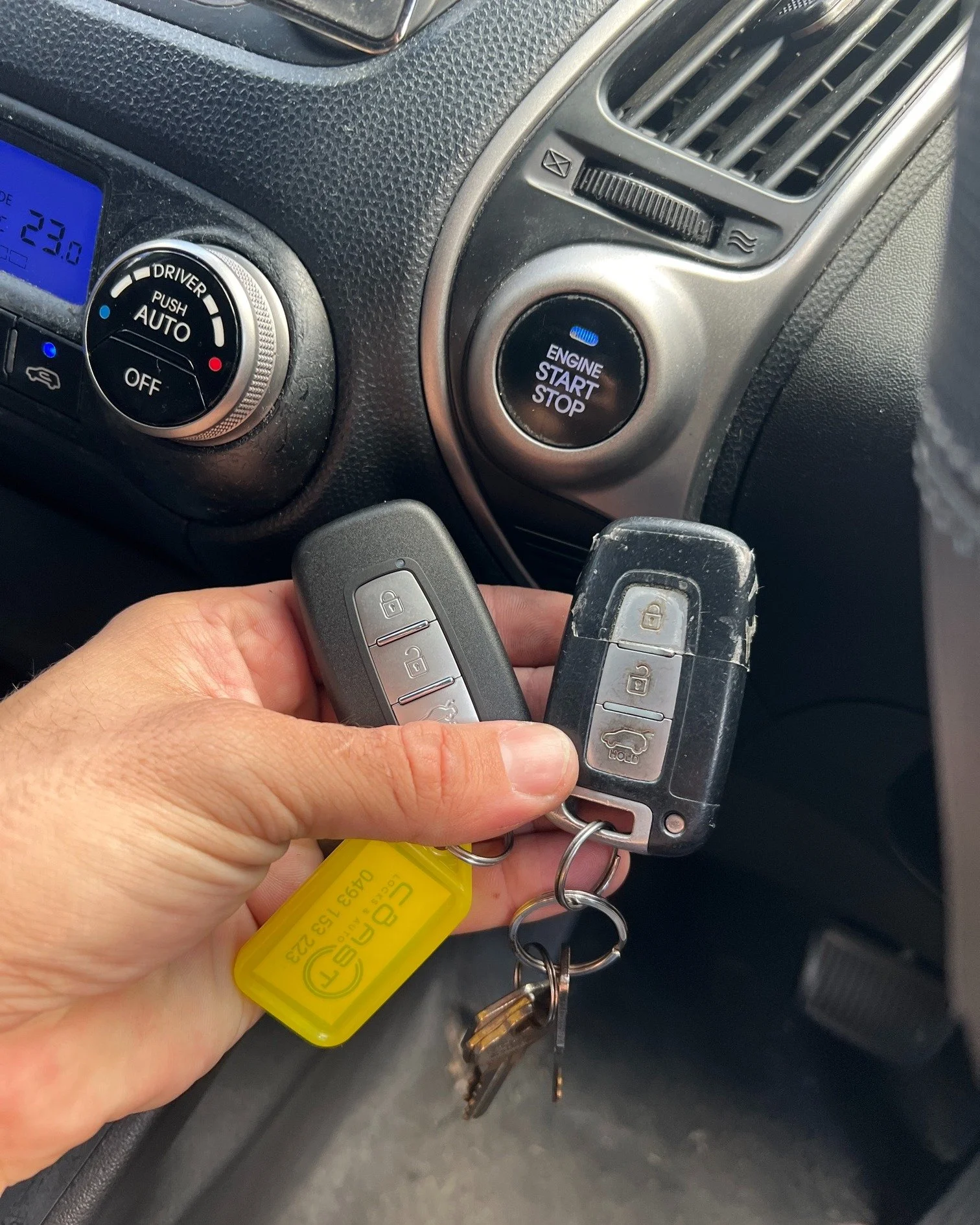 Person holding car keys inside a vehicle with a visible dashboard, climate control knob, and engine start/stop button.