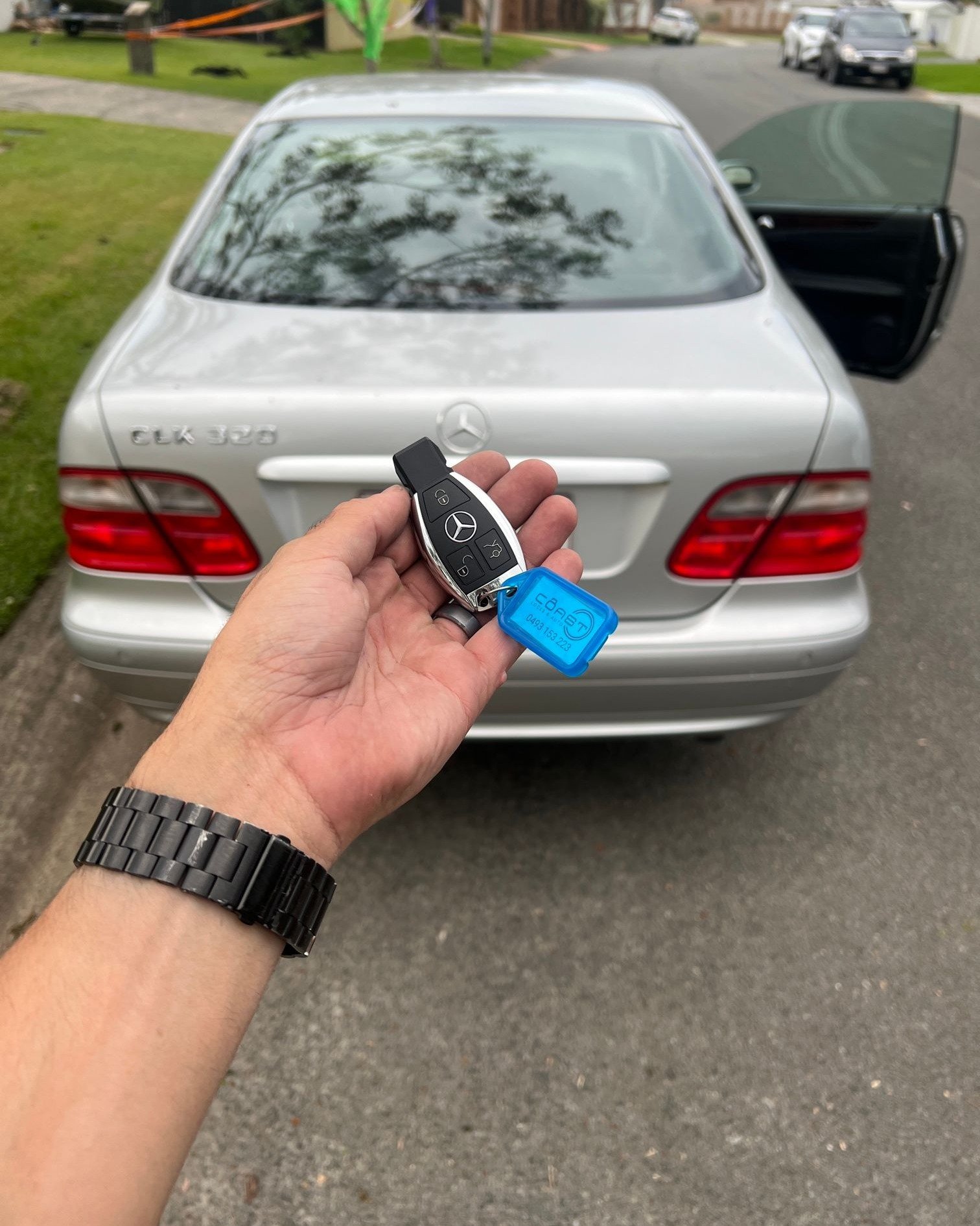 A person holding a Mercedes-Benz key fob in front of a silver Mercedes-Benz car parked on a residential street.
