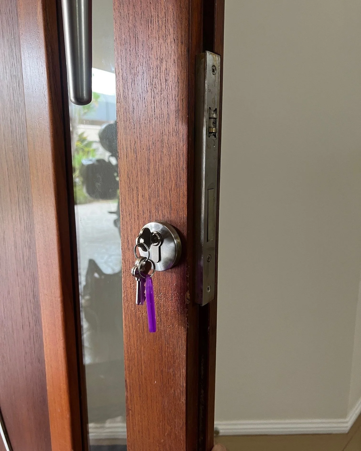 Close-up of a wooden door with a metal lock and keys hanging from the lock. The door is slightly open, revealing an outdoor area with plants and a person holding a camera reflected in the glass.