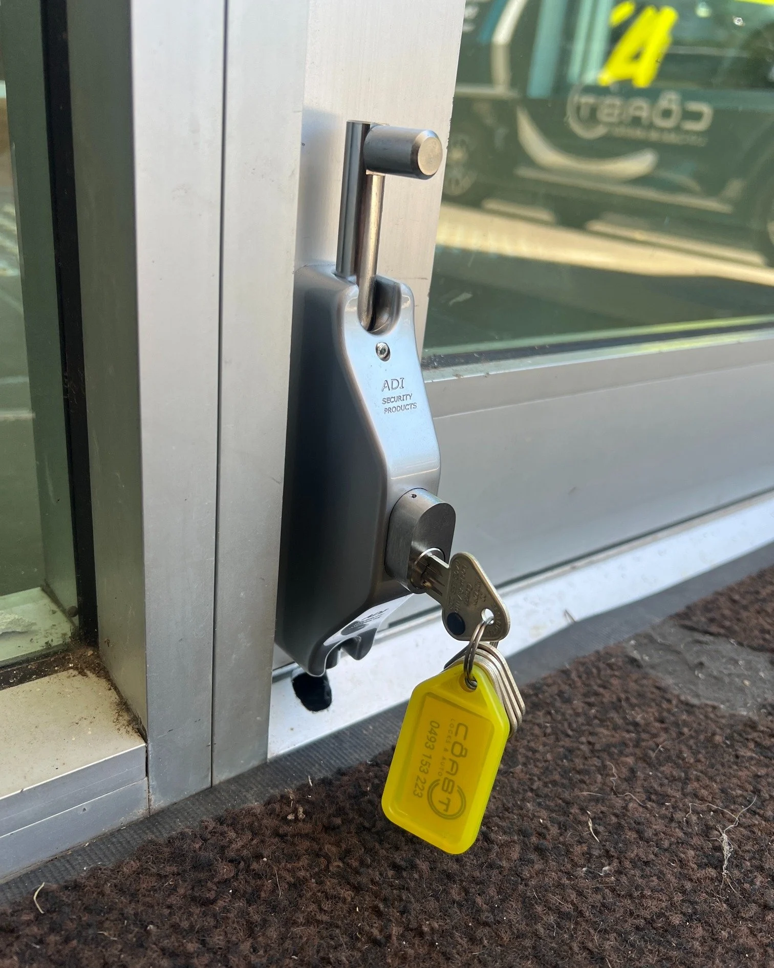Close-up of a silver door lock with a key inserted, attached to a glass door frame, with a yellow key tag hanging from the key.