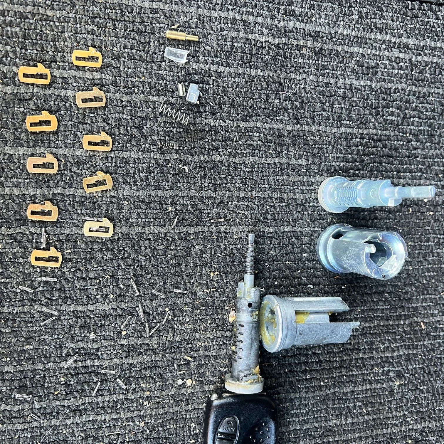 Disassembled automotive ignition lock cylinder with associated small parts, screws, and a key on a dark textured surface.