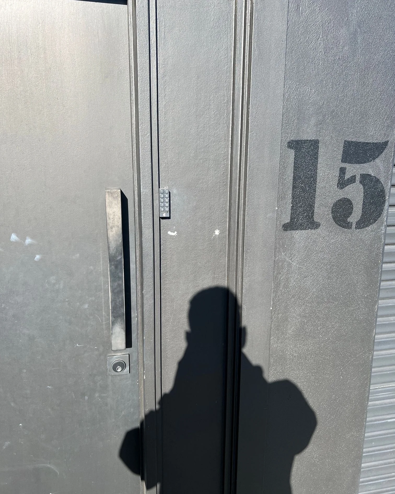 Gray metal storage locker with the number 15 painted on it, a keypad lock, and a shadow of a person taking the photo.