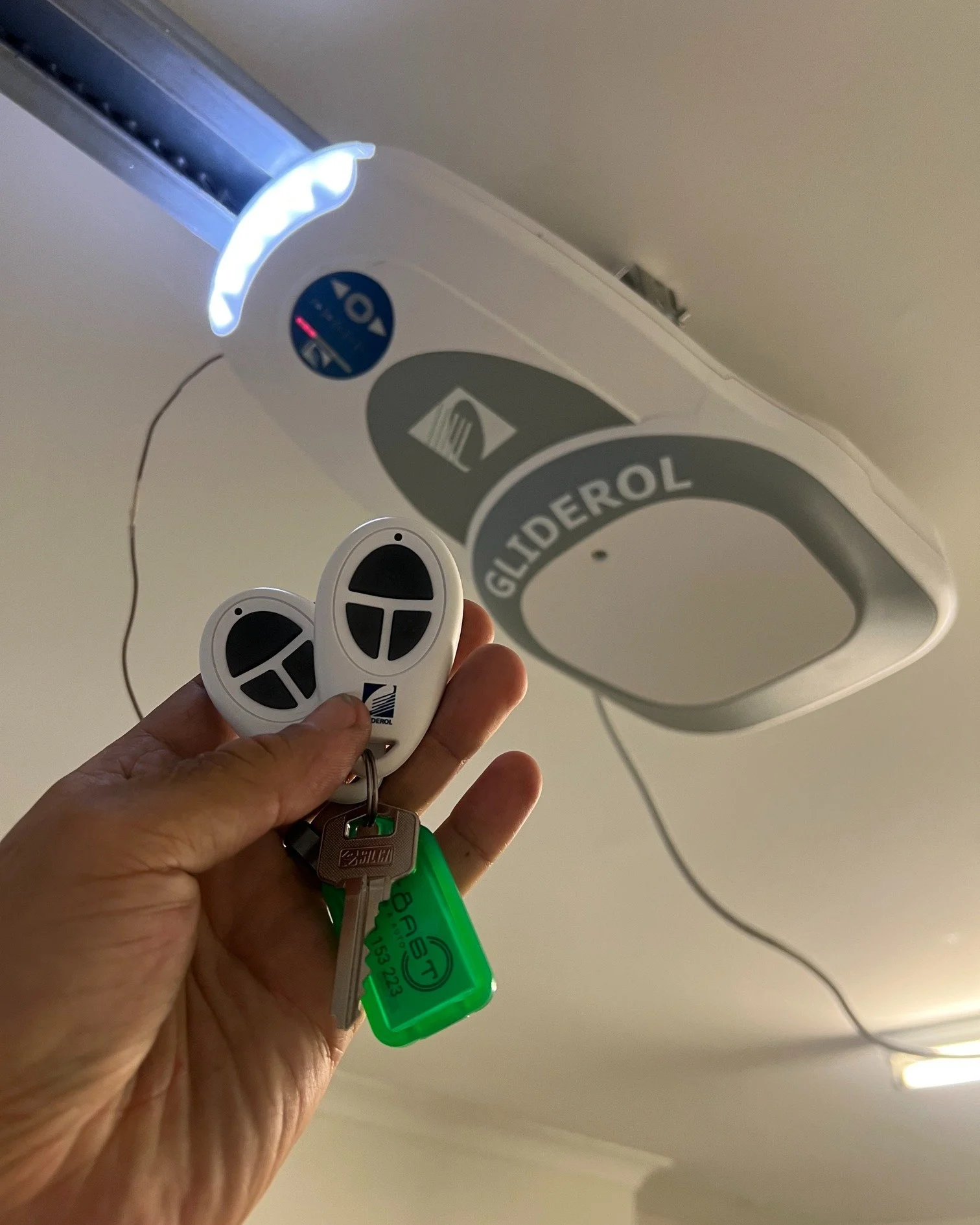 Person holding remote controls and keys near an automatic garage door opener labeled 'Gliderol'.