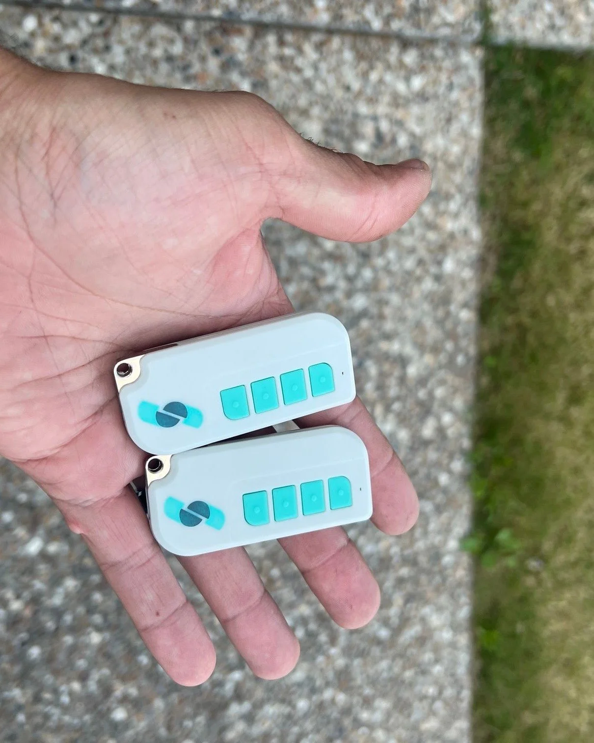 A person holding two small portable electronic devices with blue buttons and a logo, outdoors on a gravel path with grass on the side.