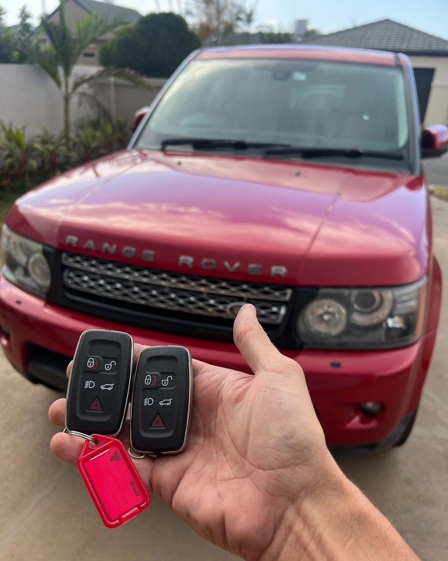 A person holding two car key fobs in front of a red Range Rover parked in a driveway with houses and landscaping in the background.