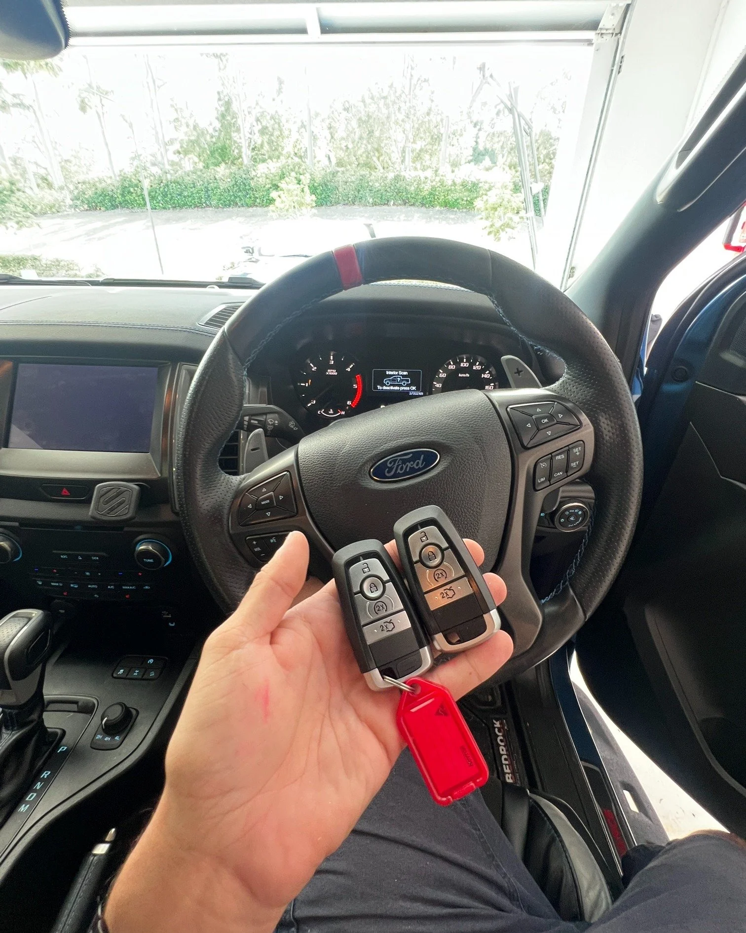 Inside a vehicle, a person holds two Ford car key fobs with a red key tag near the steering wheel, which has the Ford logo. The dashboard and control panel of the vehicle are visible, with a view through the windshield showing some greenery and trees outside.
