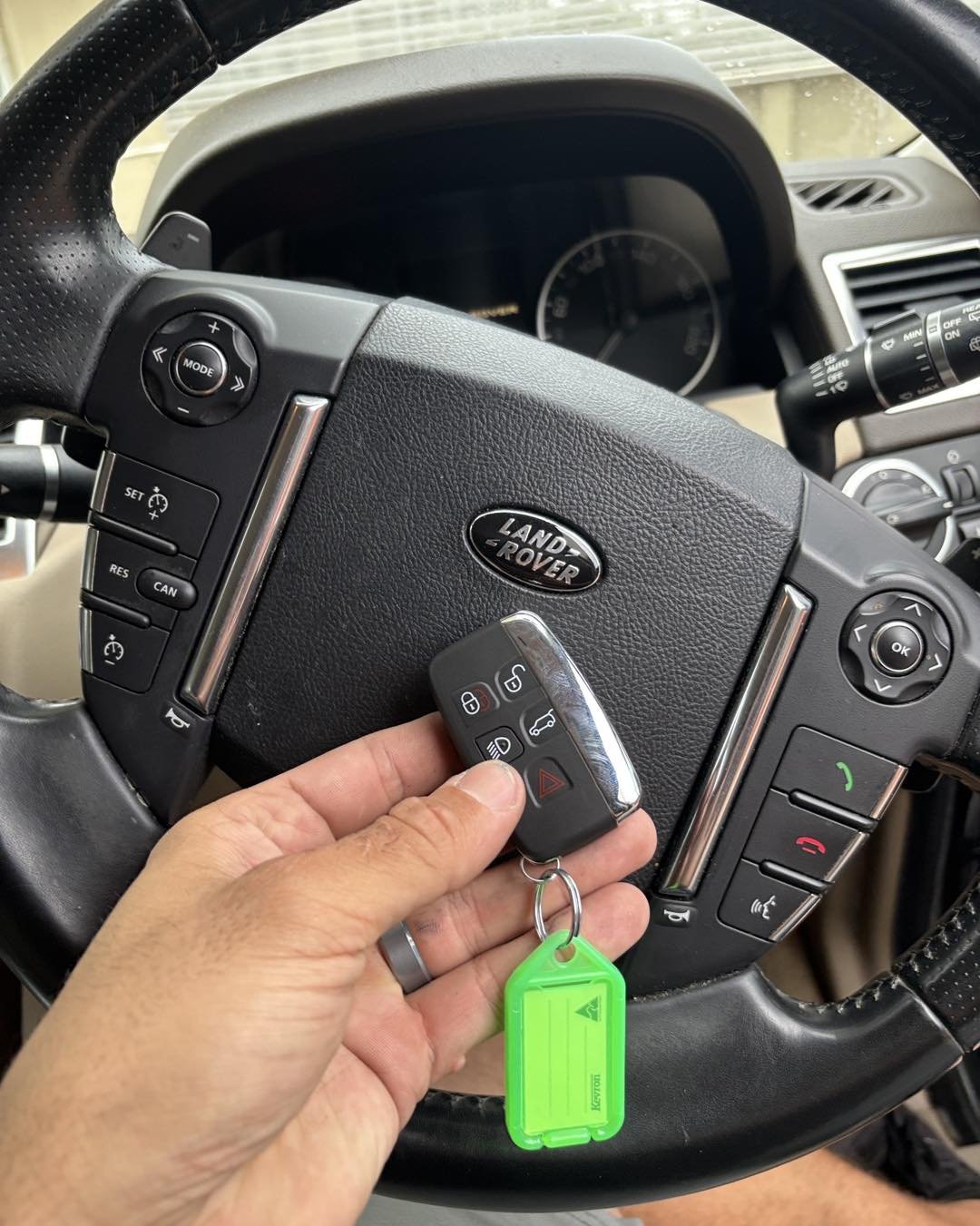 Inside a Land Rover vehicle, a hand holds a car key fob with buttons for lock, unlock, trunk, and alarm, in front of the steering wheel.