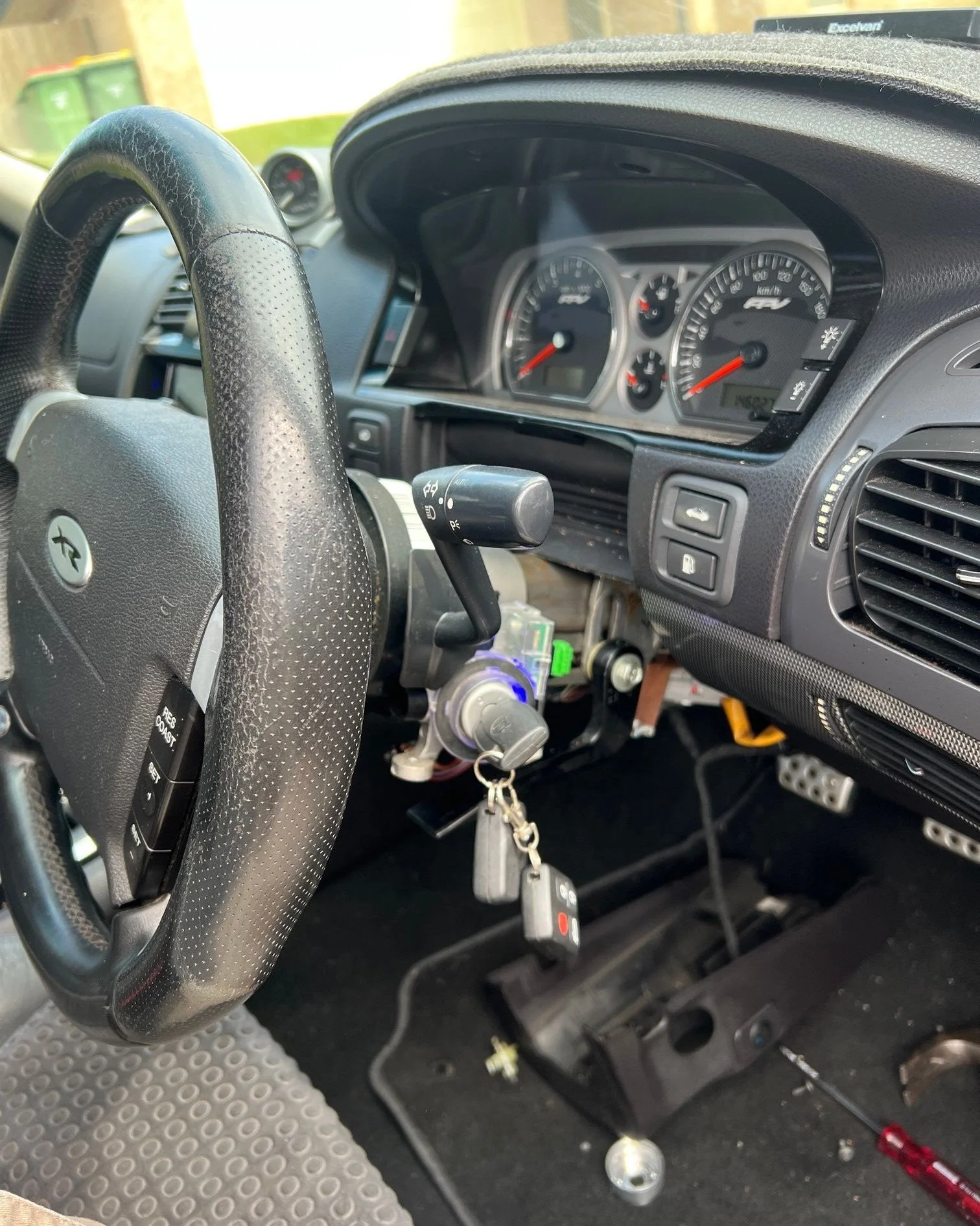 Inside view of a car dashboard showing a steering wheel, speedometer, tachometer, and various controls. The ignition is on with keys inserted, and there is a device attached to the dashboard with visible wires. The car's footrest and pedals are also visible.