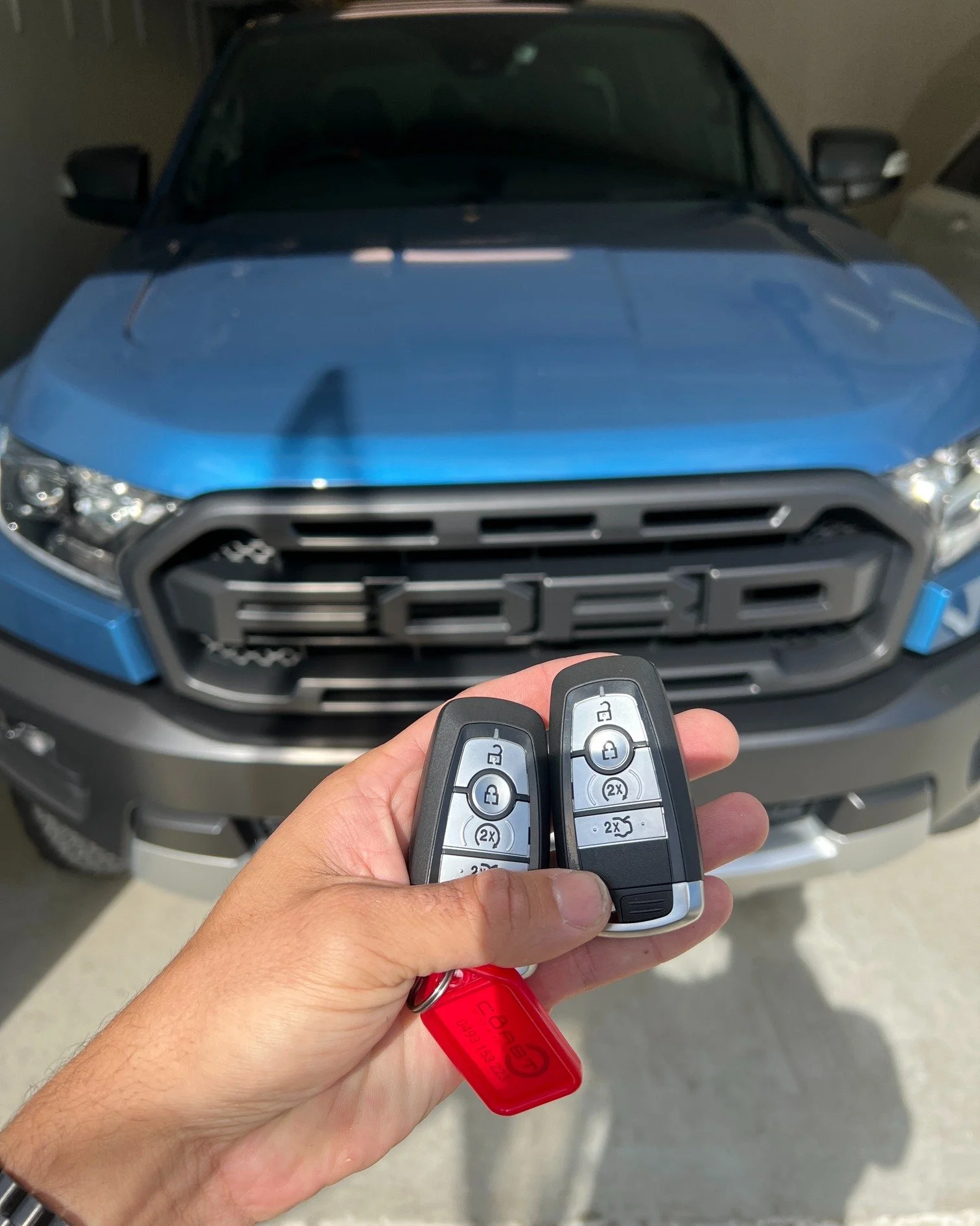 A person's hand holding two Ford car key fobs with a blue Ford vehicle in the background.