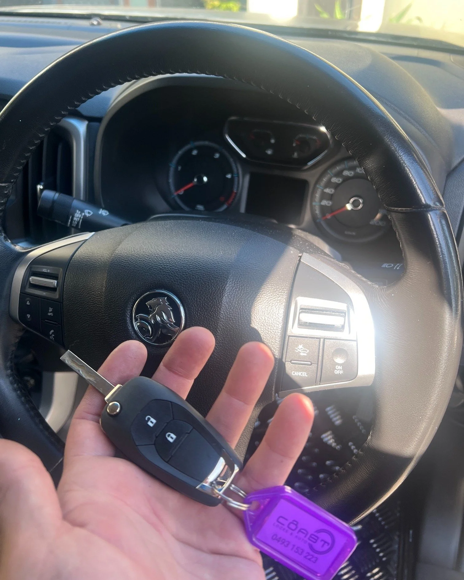 Interior of a car showing a steering wheel with a person's hand holding car keys in front, with visible buttons and a purple key tag. The dashboard and instrument gauges are also visible.