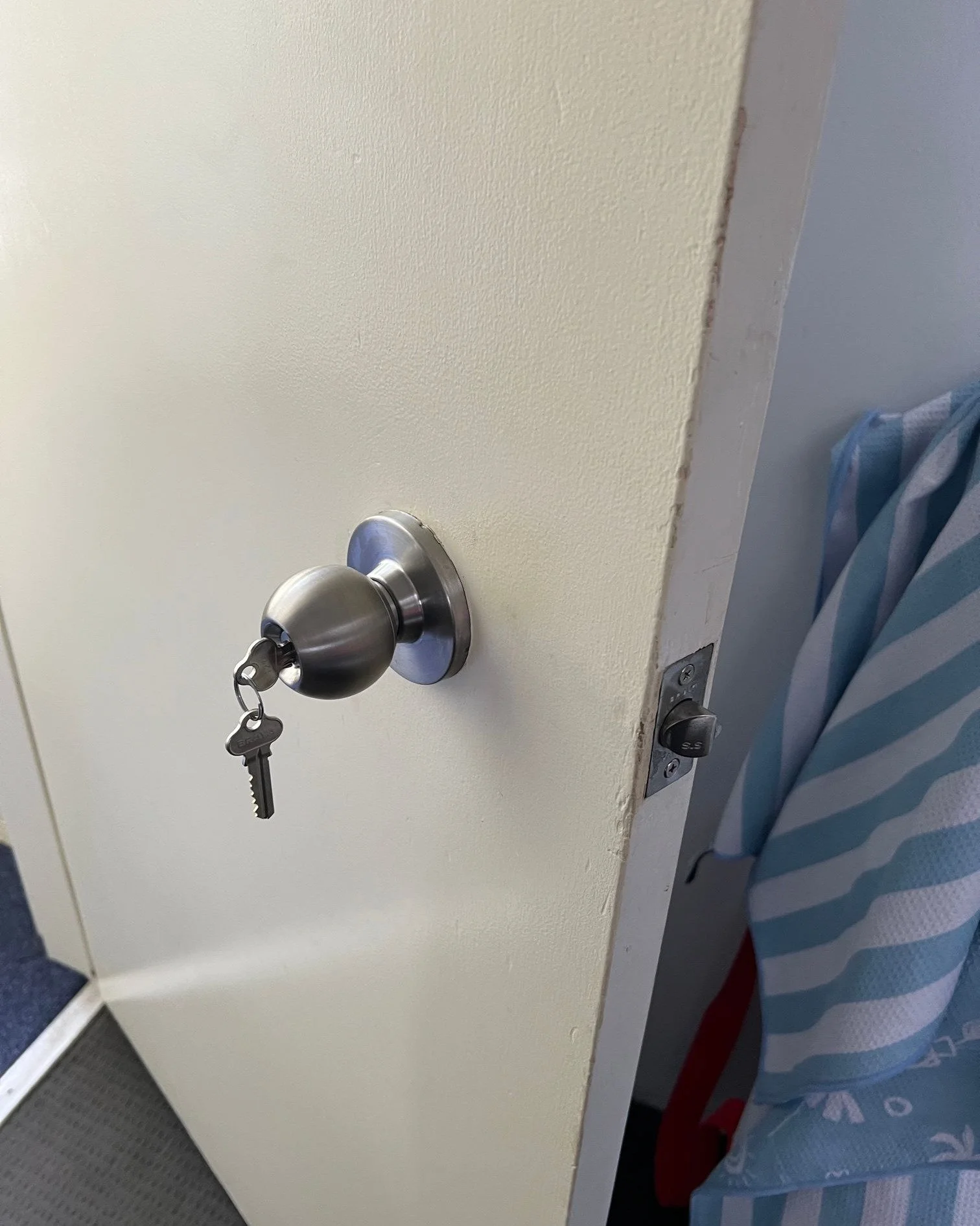 A white door with a metal doorknob and a key inserted into the lock. Behind the door, there is a glimpse of a blue and white striped curtain or fabric.
