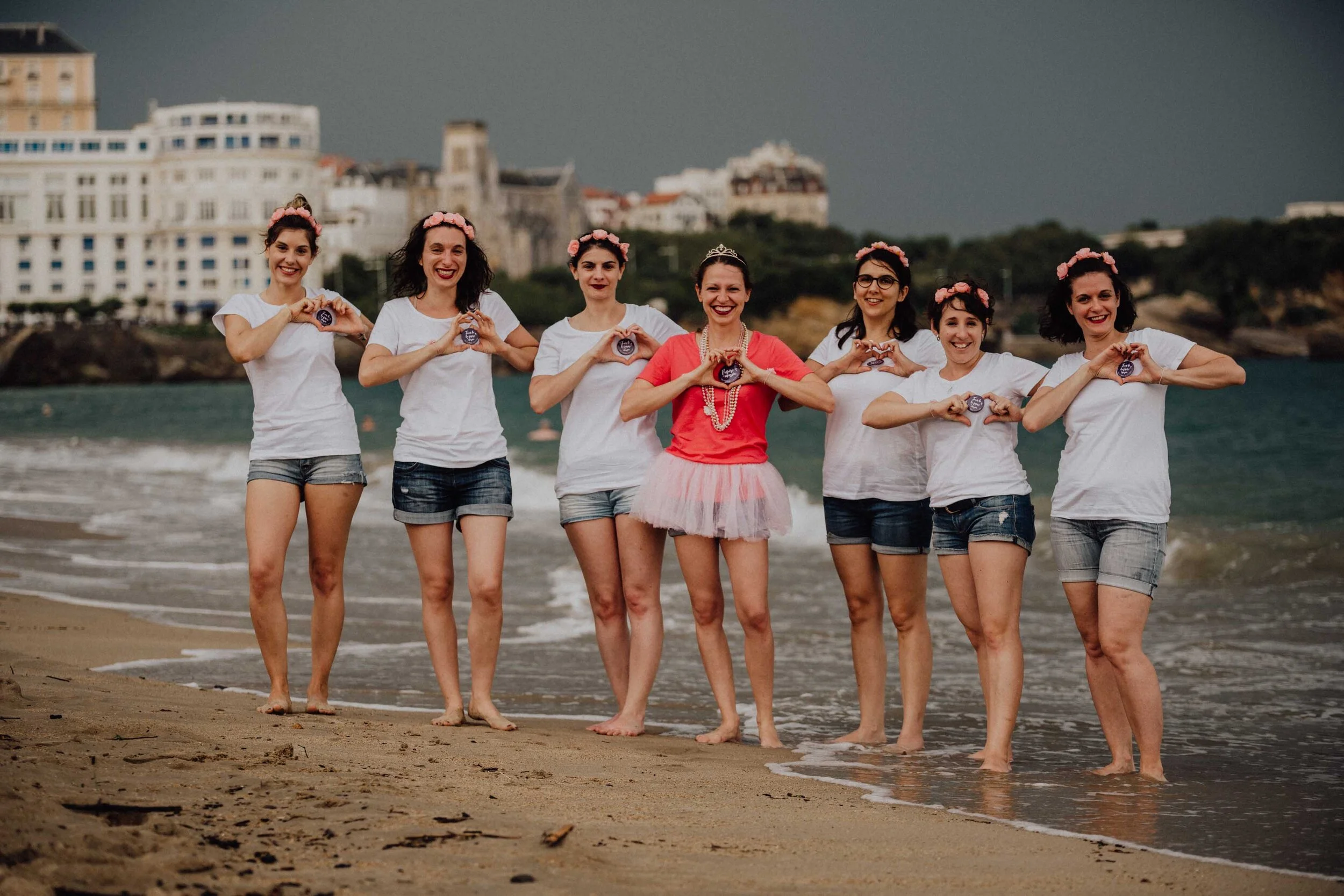Groupe de sept femmes souriantes se tenant par la main sur une plage, portant des couronnes de fleurs roses et des vêtements décontractés. Arrière-plan de bâtiments et ciel orageux.