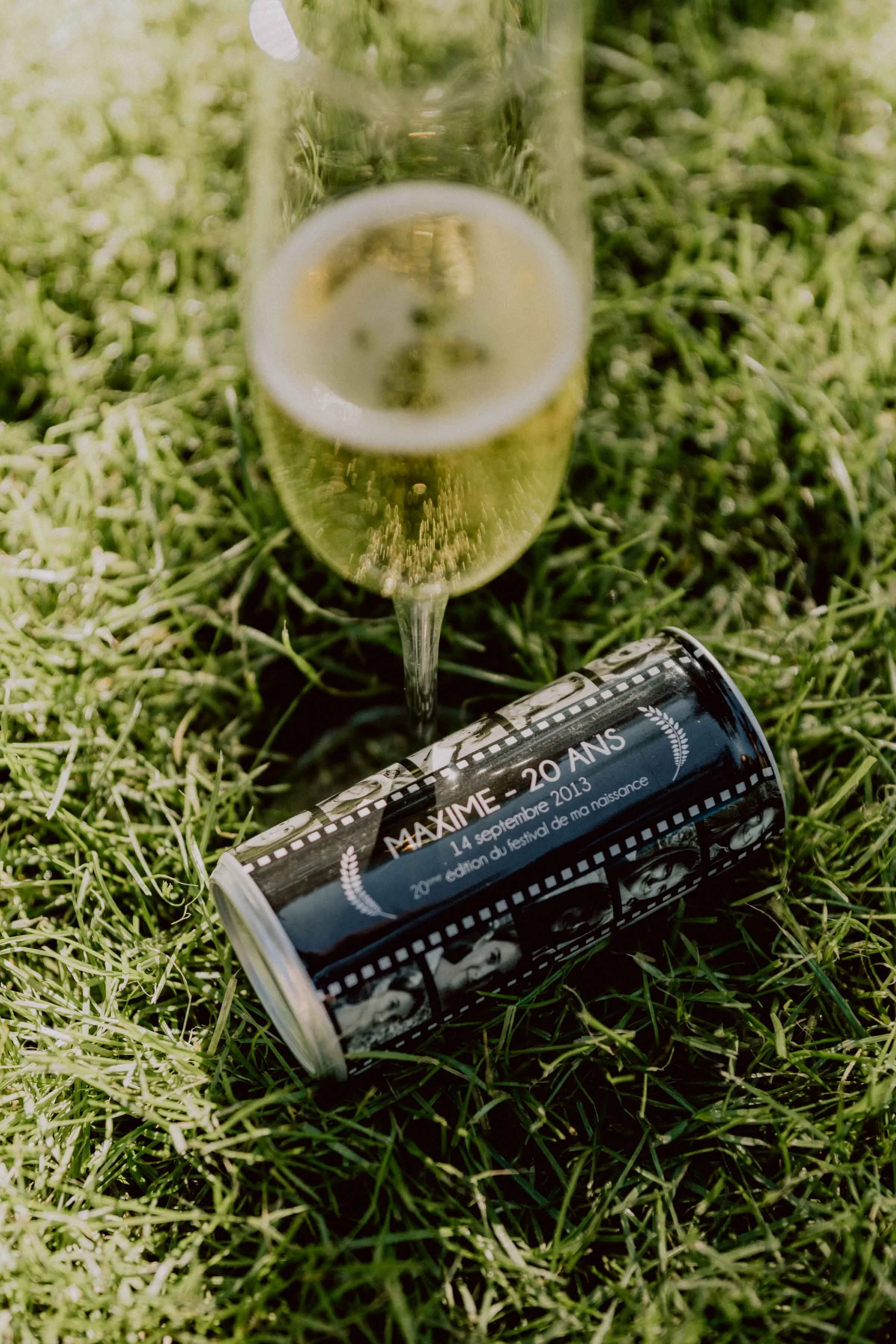 Une bouteille en verre de champagne repose sur l'herbe, avec un verre de champagne à moitié rempli à côté. Il y a aussi une canette métallique sur le sol, avec des informations en français sur une fête ou un anniversaire.