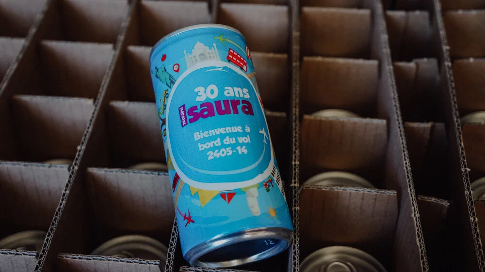 Une canette de boisson en aluminium avec une étiquette bleu, célébrant le 30e anniversaire de Laura, avec une inscription en français. La canette est placée dans une boîte en carton contenant d'autres canettes.