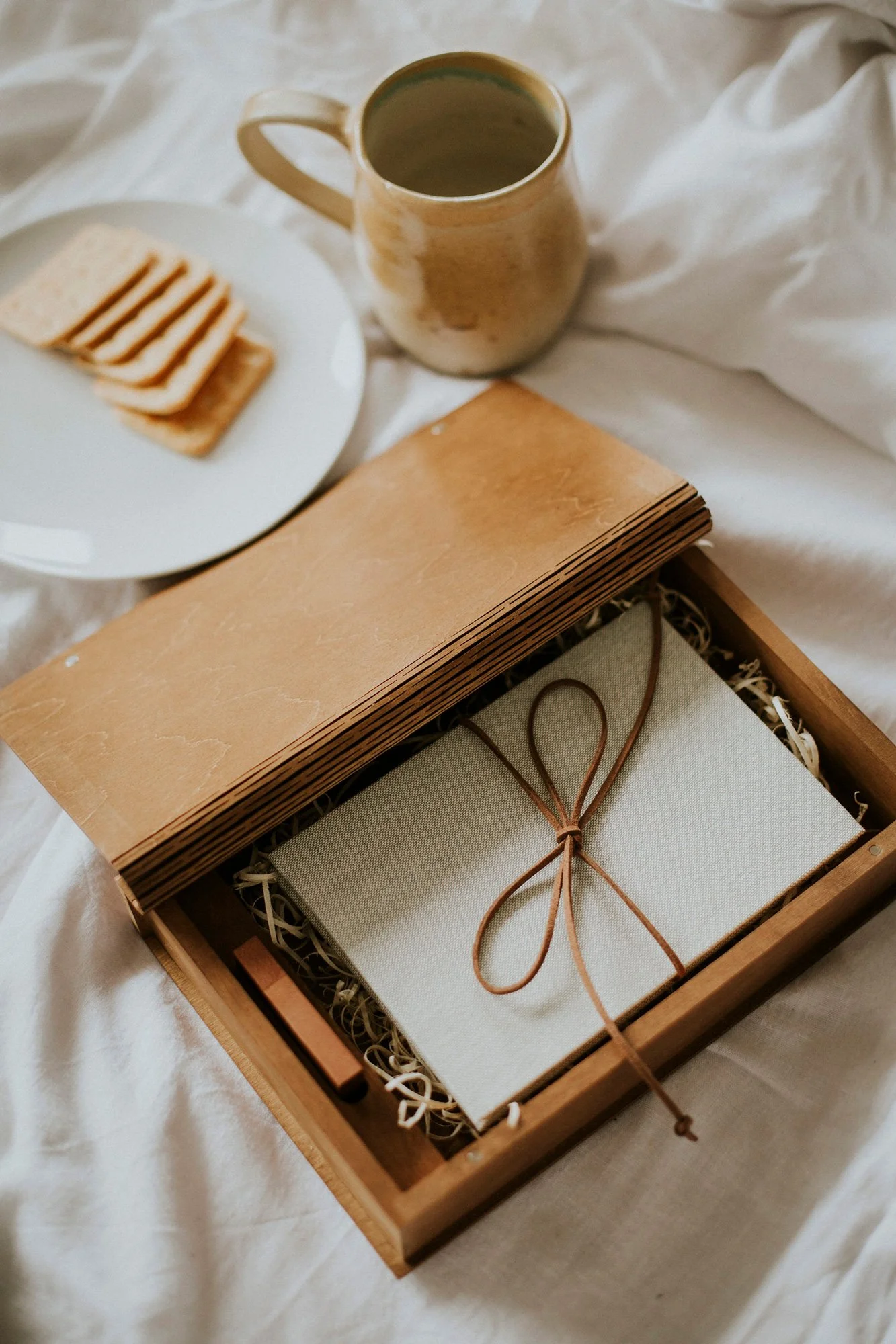 Une boîte en bois contenant un cadeau emballé avec un ruban en cuir, une tasse de café et des biscuits sur une surface en tissu blanc.