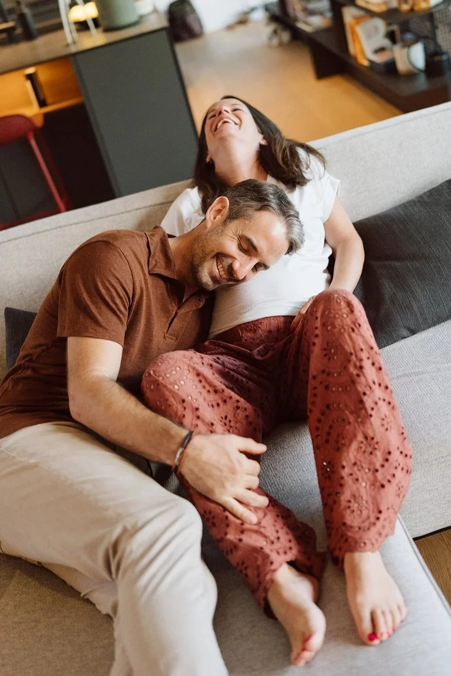 Séance photo grossesse à domicile : un couple éclate de rire sur le canapé, capturant un instant spontané, joyeux et complice dans l’attente de leur bébé.
